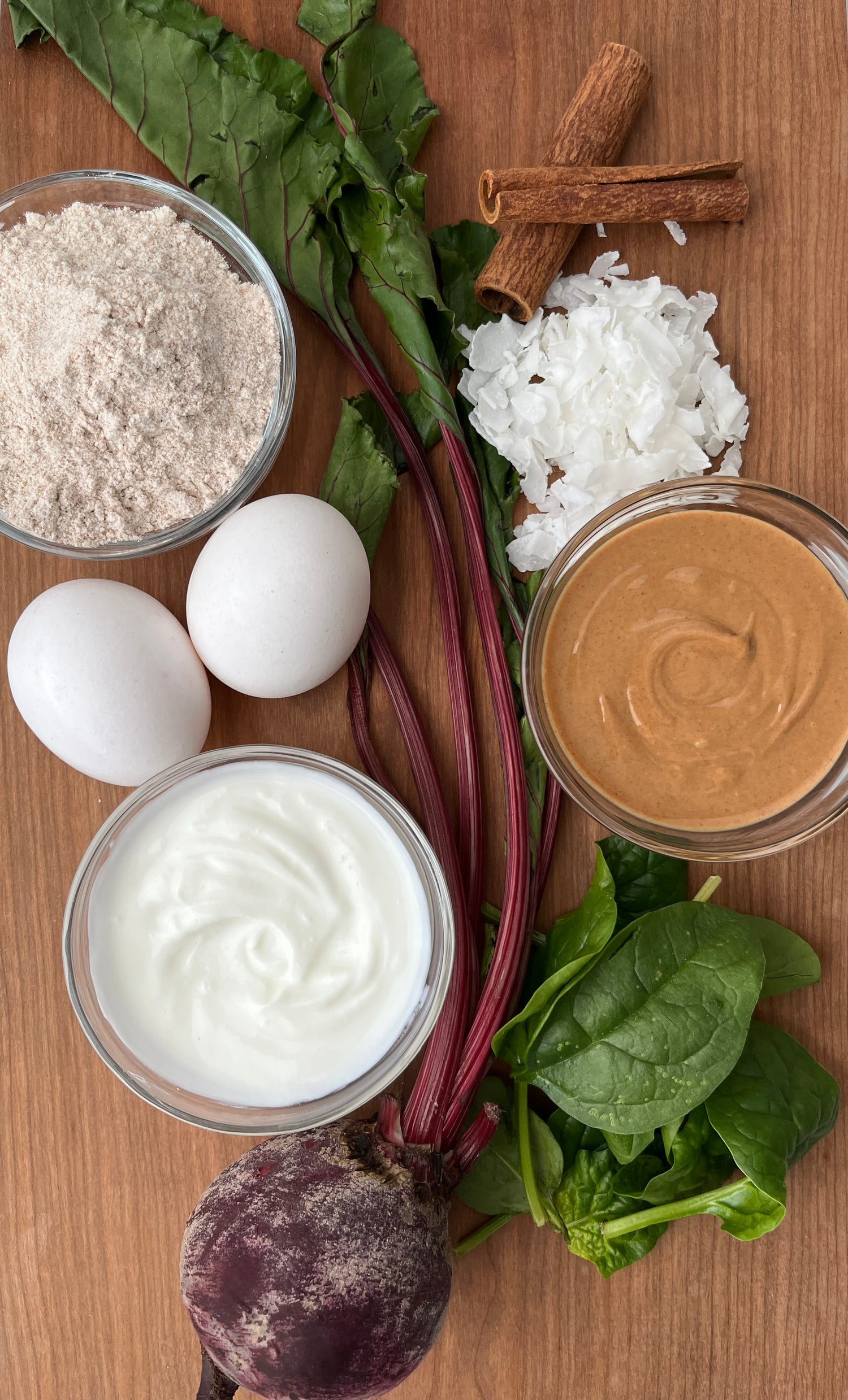 Picture of a beet, eggs, cinnamon sticks, spinach, coconut flakes, and bowls of peanut butter, yogurt, and wheat flour on a cutting board.